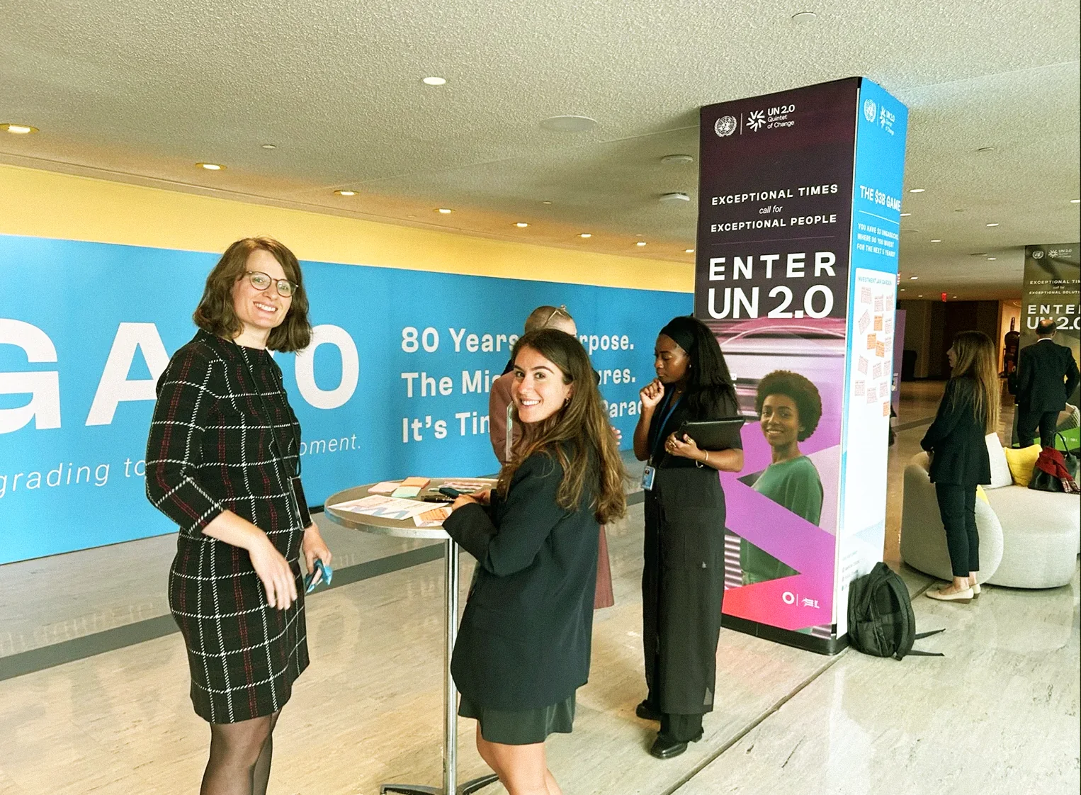 Visitors gathered at the table beside a column, with the #UNGA80 wall behind them
